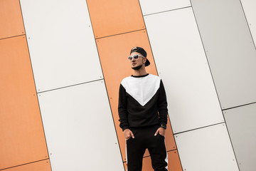 Wall Mural - Fashionable young man in a stylish sporty black suit in a trendy cap with sunglasses stands near a modern building in the city on a summer day. Handsome guy with a beard outdoors. Youth street style.