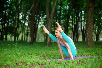 Thin brunette girl plays sports and performs beautiful and sophisticated yoga poses in a summer park. Green lush forest on the background. Woman doing exercises on a yoga mat