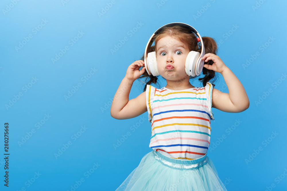 Happy smiling child enjoys listens to music in headphones over colorful bleu background. Vivid and fun emotions, happy child with pleasure listens to songs in wireless headphones - obrazy, fototapety, plakaty 
