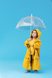 © oes - Happy funny child with transparent umbrella posing on blue studio background. Girl is wearing yellow raincoat and rubber boots. Holds a vintage travel suitcase