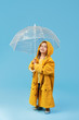 © oes - Happy funny child with transparent umbrella posing on blue studio background. Girl is wearing yellow raincoat and rubber boots. Holds a vintage travel suitcase