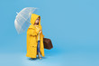 © oes - Happy funny child with transparent umbrella posing on blue studio background. Girl is wearing yellow raincoat and rubber boots. Holds a vintage travel suitcase. Space for text on right side