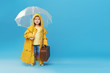 © oes - Happy funny child with transparent umbrella posing on blue studio background. Girl is wearing yellow raincoat and rubber boots. Holds a vintage travel suitcase