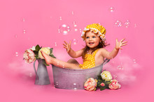 Cute Girl In A Yellow Jumpsuit And In A Yellow Hat With Sitting In A Baby Bath. Soap Bubbles On A Pink Background