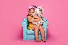 Little Girl With A Dog. A Cute Little Girl With A Dog Is Sitting In A Blue Armchair. Pink Background