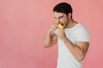 Wall Mural - hungry muscular man in white t-shirt eating banana isolated on pink