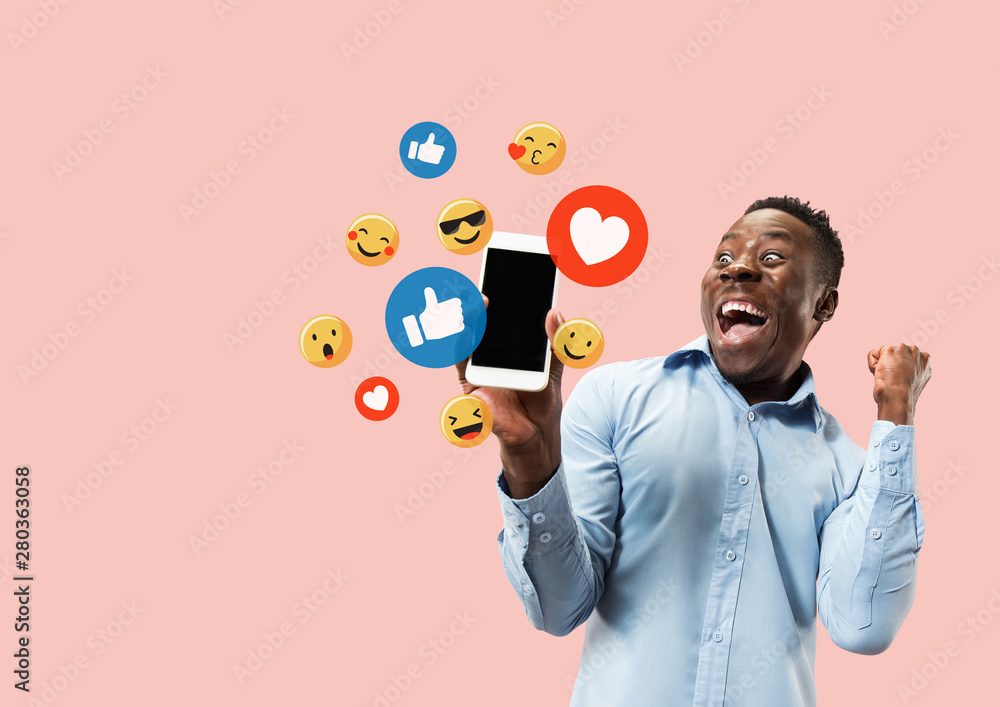 Social media interactions on mobile phone. Internet digital marketing, Chating, commenting, liking. Smiles and icons above smartphone screen, that holding by young man on pink studio background. - obrazy, fototapety, plakaty 