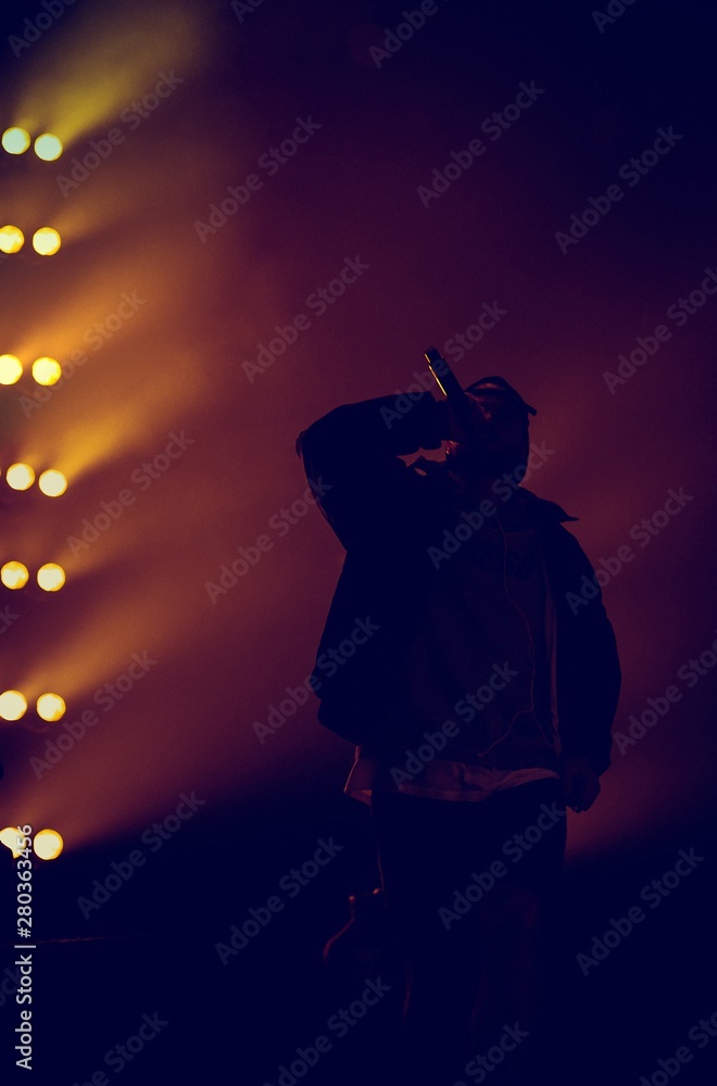 Hip hop singer with mic in hand singing on stage.Rap singer performing live on concert scene.Silhouette of rapper musician singing in radio microphone at hip hop music festival - obrazy, fototapety, plakaty 