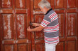 © luciano - Senior man with white beard and hair tries to open an old red door closed by lock. spanish home with red windows and doors made by wood. Rustic style. Sunlight in summer time