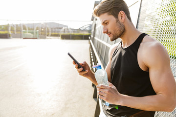 Wall Mural - Photo of young man holding cellphone while standing on sports ground during morning workout at green park