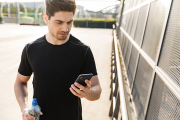 Canvas Print - Portrait of young man holding cellphone and water bottle while standing on sports ground during morning workout outdoors