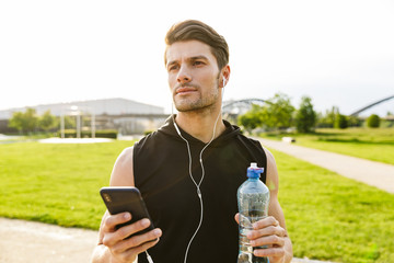 Wall Mural - Image of attractive man drinking water and holding cellphone while running with earphones at green park outdoors