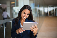 Cheerful Happy Office Worker Using Tablet In Office Hall. Young Latin Business Woman Leaning On Glass Wall And Reading On Screen. Tablet Computer Concept