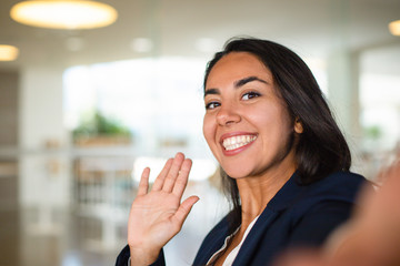 Wall Mural - Happy young woman waving hand. Close-up view of beautiful cheerful young businesswoman smiling at camera and waving hand. Emotion concept