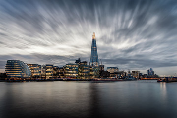 Fototapete - Die markante Skyline von London an der Themse mit den beleuchteten Wolkenkratzern am Abend mit Wolken, Großbritannien