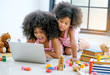 © narong - Two young African girls play with notebook computer among toys, doll and book in front of glass window.