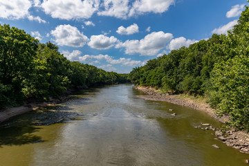 water flowing along rocky riverbank of sangamon river surrounded by trees on a sunny day with clouds