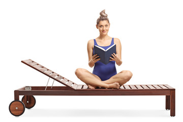 Wall Mural - Young female in a swimming suit sitting on a lounge chair with a book and looking at the camera