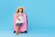 © oes - Happy child girl tourist packs clothes into a suitcase for travel, summer vacation. Toddler kid in straw hat  holding a pink suitcase on a blue background