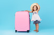 © oes - Happy child girl tourist packs clothes into a suitcase for travel, summer vacation. Toddler kid in straw hat  holding a pink suitcase on a blue background