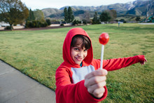 Boy In A Red Hoodie Holds Up His Red Lollipop While Outside At A Park