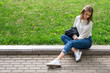 © Денис Бухлаев - beautiful slender girl is resting in city park. Caucasian girl posing sitting on curb sidewalk. Lifestyle, pullover, jeans, sneakers, jacket. Cool posing while sitting on grass in trendy look.