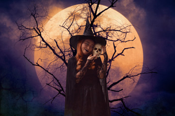 Halloween witch holding a skull standing over dead tree, full moon and spooky cloudy sky, Halloween mystery concept