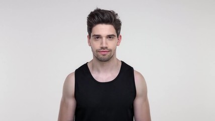 Wall Mural - Smiling young bearded sporty man in black shirt winking to the camera over gray background isolated