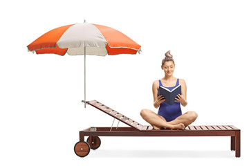 Wall Mural - Young woman in a swimming suit sitting on a sunbed under umbrella and reading a book