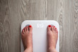 © AllUneed - Woman stands on white modern electronic scales, which show 64.5 kg. Only feet are visible. Scales stand on gray wooden floor. Space for text. Healthy lifestyle, diet, weight loss concept. Horizontal.