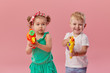 © oes - Happy little toddler boy and girl  in summer clothes holds toy water guns isolated on pink wall background. Children studio portrait. People childhood lifestyle concept.