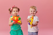 © oes - Happy little toddler boy and girl  in summer clothes holds toy water guns isolated on pink wall background. Children studio portrait. People childhood lifestyle concept.