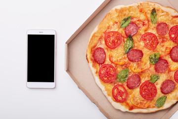 Pizza in box and smartphone with blank screen on white table, top view of delicious pepperoni isolated over light background. Modern mobile phone near box with pizza. Copy space for advertisment.