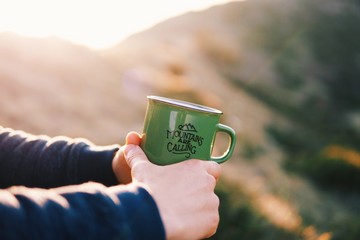 Close up shot of the tourist hands holding green mug with hot drink, traveler holds in hands a mug with hot tea, enjoying the sunset in the mountains, lifestyle relax trekking concept