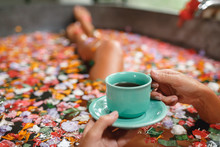 Woman In Bath With Petals With Cup Of Herbal Tea. Young Woman Relaxing In Bath With Petals In Tropics. Hand With Cup Of Tea Closeup