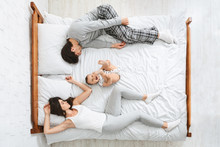 Exhausted Parents Sleeping On Sides Of Bed, Active Baby Playing In Middle
