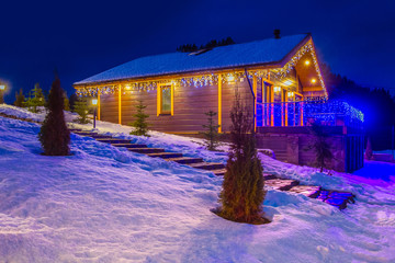 Wall Mural - Christmas Eve. The house is decorated with garlands. Christmas holidays. Wooden house in the illumination. Winter evening. The cottage is covered with snow. Buy a house in Canada.