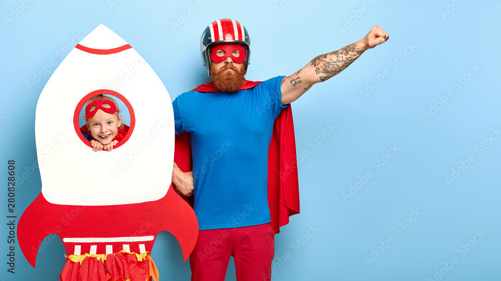 Serious bearded father raises arm, pretends being superhero, wears helmet, mask, cape and blue t shirt, plays with little daughter who looks through hole of paper rocket. My dad is superman! - obrazy, fototapety, plakaty 