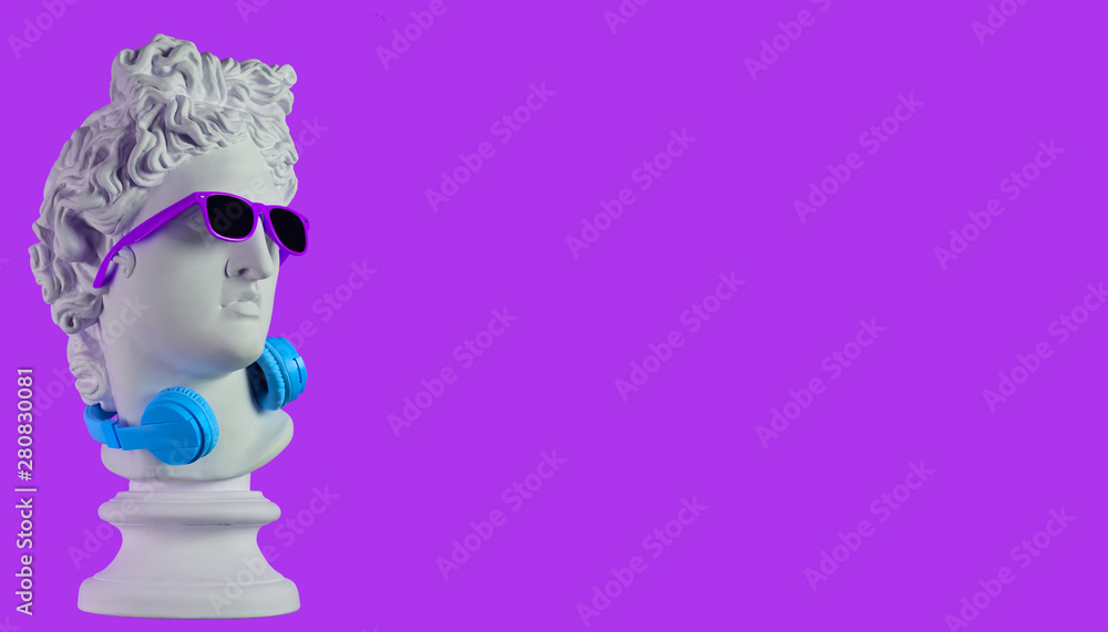 Statue. Earphone on a purple background. Gypsum statue of David's head. Creative. Plaster statue of Apollo's head in blue sunglasses. Minimal concept art. - obrazy, fototapety, plakaty 