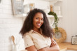© Anatoliy Karlyuk - Horizontal shot of beautiful young black woman in beige shirt crossing arms on chest, looking at camera with confident toothy smile, posing indoors against stylish kitchen interior background