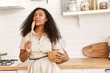 © Anatoliy Karlyuk - Attractive young African American housewife in beige dress standing in kitchen with utensils and wooden spoon having pensive facial expression, thinking what to cook for dinner. Cuisine and food