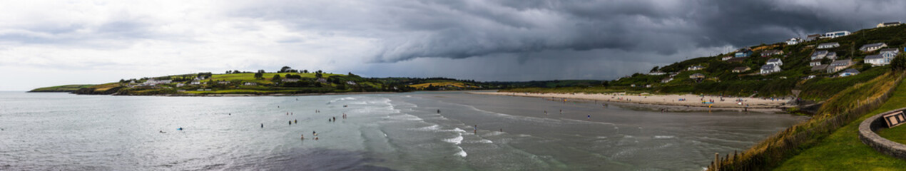 Wall Mural - A panoramic view of the Inchydoney beach on an overcast day with people surfing, swimming, and enjoying the warm weather