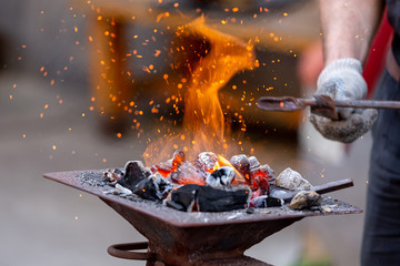 Closeup of blacksmith hand holding tongs with metal piece and heating it in fire before forging, selective focus. Blacksmithing concept.