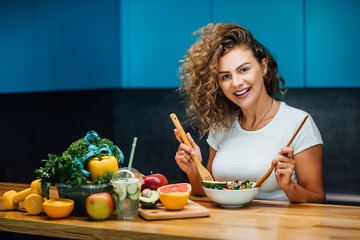 Young and happy woman eating healthy salad sitting on the table with green fresh ingredients indoor.