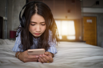 Wall Mural - Young girl listening music from smartphone on bed at home. Asian woman lying on bed holding mobile phone listening music with headphones in bedroom while text message phone.