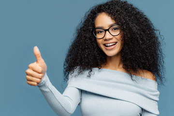 Wall Mural - Horizontal shot of lovely dark skinned woman gives thumb up, wears fashionable sweater, eyewear, demonstrates approval, isolated on blue background. Nice choice, good job, well done concept.