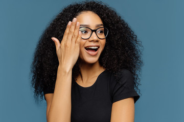 Wall Mural - Headshot of beautiful curly woman looks aside, touches face, wears transparent glasses, black casual t shirt, isolated on blue background, notices something pleasant. People and positiveness concept