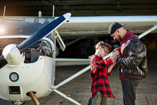 Little Boy In Aviator Glasses And His Big Brother In Black Pilot Jacket Stand Near White Propeller Airplane, Assemble Toy Plane Together, Both Wearing Red Plaid Shirts And Jeans. Outdoor Shot.