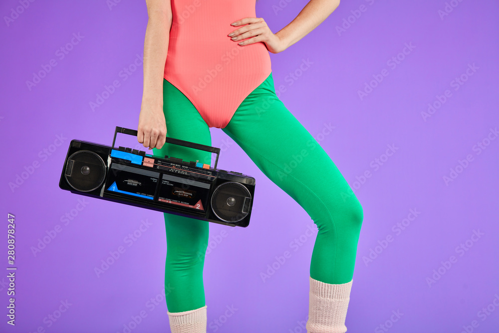 fitness girl stands in comfortable pink bodysuit and green leggins with one hand holding portable retro audio player and other is on her waist, has nice body curves and well-shaped - obrazy, fototapety, plakaty 
