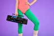 fitness girl stands in comfortable pink bodysuit and green leggins with one hand holding portable retro audio player and other is on her waist, has nice body curves and well-shaped